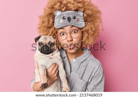 Une jeune femme surprise aux cheveux bouclés garde les lèvres repliées sur elle-même porte un chien de chien de chien de chien de chien de chien de chien de chien de chien de chevet passe du temps libre à la maison avec un animal de compagnie portant un pyjama et un masque de nuit isolé sur fond rose. concept Animaux