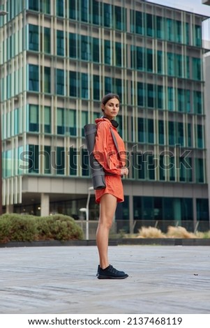 Full length shot of sporty muscular woman dressed in anorak and shorts carries rolled karemat for fitness mat looks seriously at camera poses against urban building. Healthy lifestyle concept