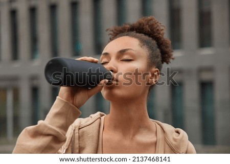 Horizontal shot of sporty woman drinks refreshing water from bottle keeps eyes closed feels thirsty after workout wears sweatshirt leads healthy lifestyle. Tired athletic female exercises regularly