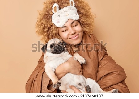 Agréable femme aux cheveux bouclés souriante portant agréablement un chien de chien de chien de chien qui dormira avec un animal de compagnie enveloppé dans une pose de couette dans une chambre isolée sur fond beige. Animaux domestiques et temps de sommeil.