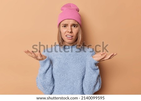 Hesitant clueless blonde woman spreads palms feels confused and bewildered frowns face wears pink hat casual blue jumper cannot make decision isolated over brown background. Human reactions.