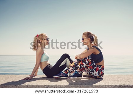 Sport fitness girls relaxing after training outdoor. Young couple talking sitting in waterfront after running and training exercise outside in summer. Caucasian and asian sports women models.