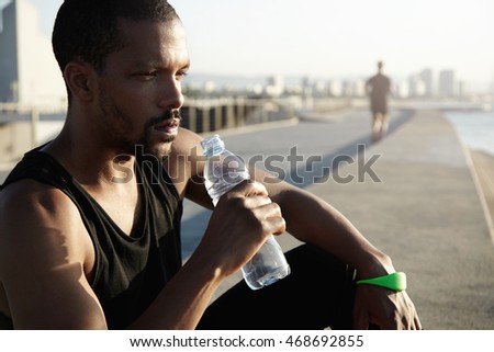Healthy lifestyle concept. Profile portrait of black sportsman with muscular body sitting on pavement in morning sun after training exercises, holding bottle, drinking water, looking into distance