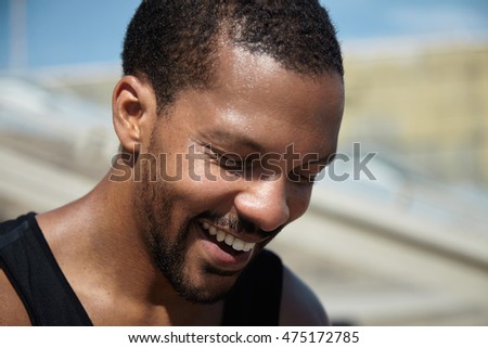 Close up shot of happy African athlete with healthy sweaty skin smiling showing his white teeth, standing against blurred urban background, relaxing after his morning run, preparing for marathon