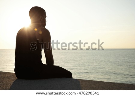 Dark-skinned young athlete having rest after exercising in open air, looking into distance, meditating, watching sunset sitting against blue sea background with copy space for your advertisement