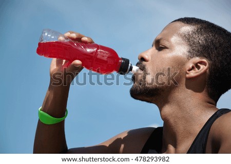 Headshot of exhausted African American athletic runner with sweaty skin wearing sleeveless black shirt, quenching his thirst with red juice or shake, relaxing after hard workout training outdoors