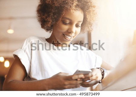 Portrait of good-looking African female student dressed casually holding mobile phone, typing messages, communicating with friends via social networks, using high-Internet connection at cafe