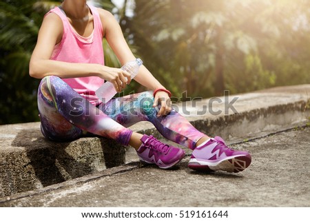 Fit woman athlete drinking water out of plastic bottle after hard running workout. Cropped shot of female runner in pink top and space print leggings resting after successful jogging exercise outdoors