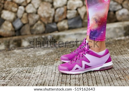 Cropped shot of fit woman runner wearing violet running shoes and space print leggings standing on stone concrete while getting ready for jogging workout. Close up of athletic pair of female legs