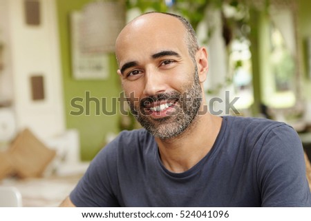 People and lifestyle concept. Happy middle-aged unshaven man wearing casual t-shirt looking at camera with cheerful smile while having lunch at outdoor cafe sitting against green interior background
