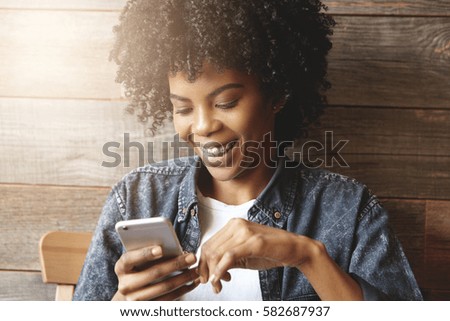 Indoor shot of dark-skinned girl with cute smile and braces enjoying free wi-fi at coffee shop, surfing internet on mobile phone, messaging friends online, inviting them to party at her place tonight