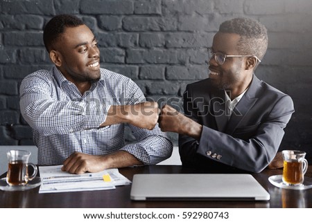 Candid shot of happy successful dark-skinned businessmen wearing formal clothing fist-bumping while cheering and congratulating each other after making profitable deal and signing good contract