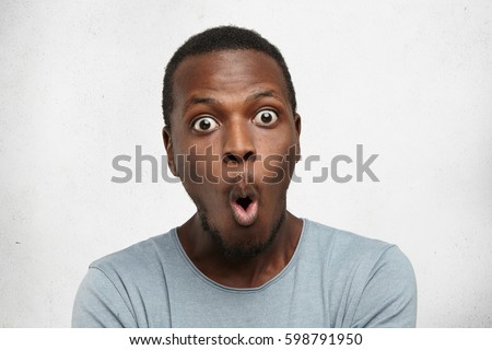 Headshot of goofy surprised bug-eyed young dark-skinned man student wearing casual grey t-shirt staring at camera with shocked look, expressing astonishment and shock, screaming 