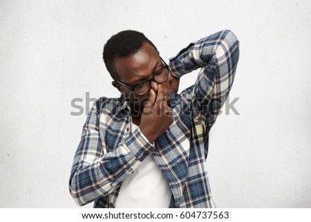 Grumpy stinky young dark-skinned male in glasses and shirt raising elbow, sniffing sweaty armpit with disgusted expression, pinching nose, can't stand unpleasant smell of sweat, posing at white wall