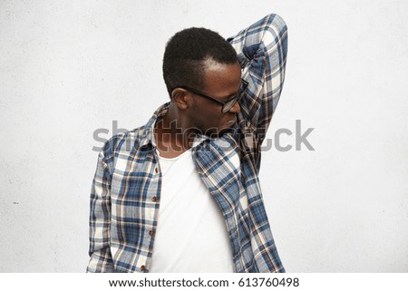 Disgusted young male wearing checkered shirt and glasses smelling wet sweaty armpit after stressful meeting, feeling nauseous, screwing lips. Black man can't stand bad smell. Hyperhidrosis and hygiene