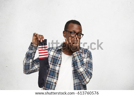 Fastidious young African American man wearing glasses and shirt over white t-shirt holding sweaty dirty stinky sock in his hand and pinching nose, his look expressing disgust with unpleasant smell