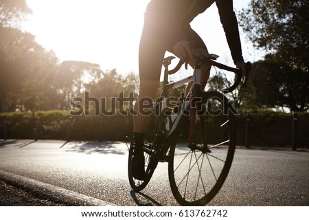 Cropped detailed shot of unrecognizable male wearing cycling clothing riding racing bike along asphalt road in urban park in warm sunny morning. Sports, fitness, extreme and active healthy lifestyle