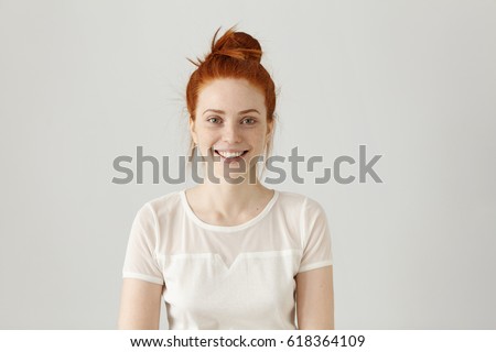 Cheerful gorgeous young woman wearing her ginger hair in knot smiling happily while receiving some positive news. Pretty girl dressed in white blouse looking at camera with excited joyful smile