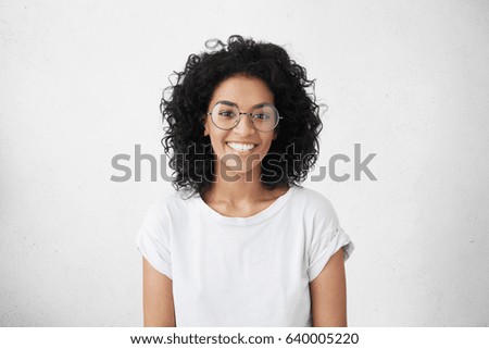 Photo of Indoor portrait of beautiful brunette young dark-skinned woman with shaggy hairstyle smiling cheerfully, showing her white teeth to camera while feeling happy and carefree on her first day-off