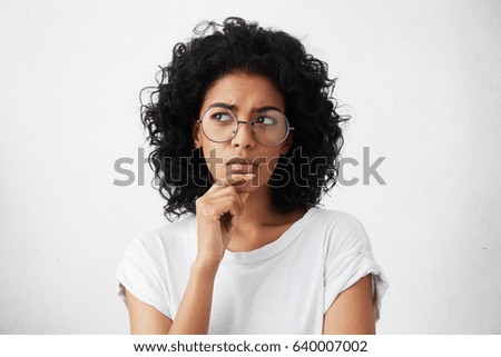 Isolated portrait of stylish young mixed race woman with dark shaggy hair touching her chin and looking sideways with doubtful and sceptical expression, suspecting her boyfriend of lying to her Stock foto © 