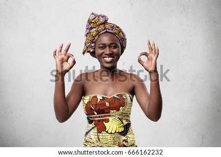 Everything is just fine! Beautiful cheerful African woman wearing bright scarf on head and elegant dress showing ok sign demonstrating her satisfaction and happiness agreeing with something.