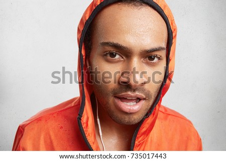 Portrait of wet dark skinned male listens to music, wears hoody, has walk on rainy day, looks confidently into camera. Young sportsman being sweaty after running, exhausted and tired. Active lfestyle