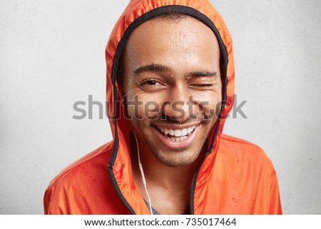 Indoor shot of cheerful positive mixed race man with healthy pure skin, being wet after going in for sport on rainy weather, blinks eye, listens favourite music, enjoys weekends. Sweaty runner in hood