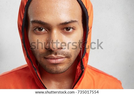 Close up portrait of attractive young man wears hood, being wet to skin after running at rainy weather, looks with appealing dark eyes directly into camera. Young sportsaman sweaty after training