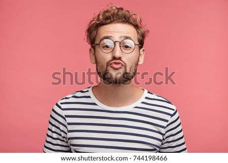 Funny male with curly trendy hairstyle, rounds lips as going to kiss someone, looks in anticipation, isolated over pink background. Fashionable bearded man epresses positive emotions, makes grimace