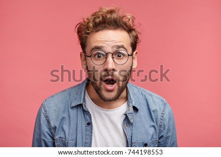 Stupefied attractive young bearded man looks with astonishment into camera, being amazed with negative news. Emotional hipster guy expresses surprisment, doesn`t believe his eyes. Facial expression