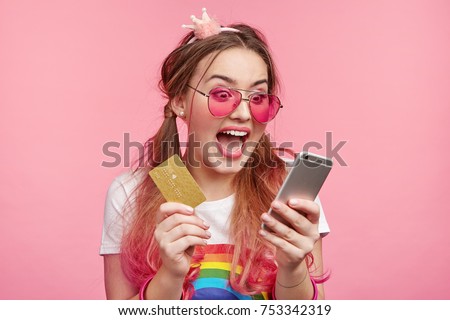 Headshot of fashionable young female looks crazy as verifies balance, happy to recieve large sum of money, uses smart phone with mobile banking application, keeps credit plastic card in hands