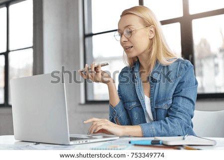 Intelligent blonde female uses voice command or recorder on smart phone in office works on laptop computer, surrounded with papers, sits at cabinet. Business, technology, communication concept