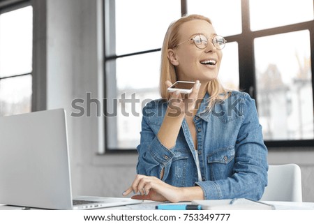 Blonde female in spectacles, wears denim shirt, uses smart phone voice recognition online, looks delightfully aside. Happy businesswoman makes voice call, sits at office table alone with modern device