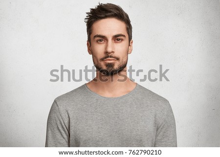 Isolated shot of young handsome male with beard, mustache and trendy hairdo, wears casual grey sweater, has serious expression as listens to interlocutor, poses in studio against white background