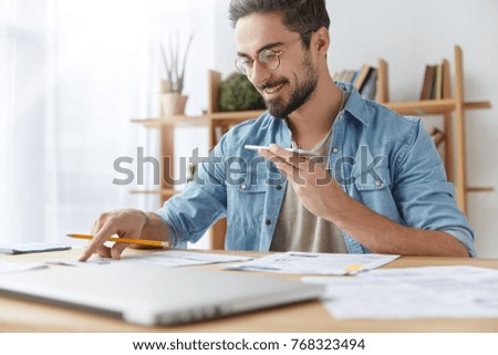 Portrait of cheerful bearded man has voice call, speaks with friend and checks documents, has happy expression. Hardworking male checks finances, does accounting, sits at desk with papers. Job concept