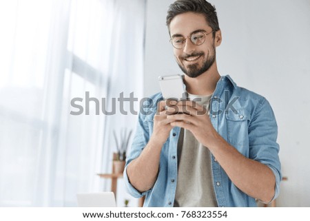 Cheerful hipster guy has thick beard and mustache, dressed casually, holds modern smart phone, glad to recieve message from best friend, reads invitation on party, rejoices good relationships.