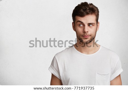 Young bearded attractive male with skeptic look, raises eyebrow in bewilderment, has some doubts and uncertainty, looks curiously aside, isolated over white background with copy space for text