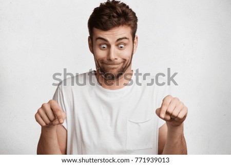 Funny surprised bearded male model looks with bugged eyes down and indicates as shows something, sees comic things, isolated over white concrete wall. Positive amazed young man poses in studio