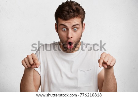 Photo of shocked bearded male looks down with great disbelief, expresses surprisment and astonishment, indicate with fore fingers at floor, isolated over white concrete wall. Caucasian man gestures