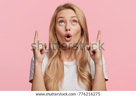 Good looking blonde female with pure healthy skin looks in amazement as indicates at something upwards, isolated over pink background. Pretty young woman sees amazing thing up, gestures indoor