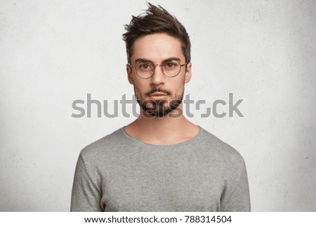 Stylish bearded male student wears round spectacles, has trendy hairstyle, looks confidently, thinks about coming session, isolated over white concrete background. People and human expressions