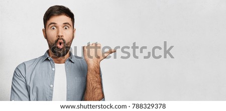 Horizontal shot of amazed young man has thick dark beard, dressed in fashionable clothes, indicates with thumb aside, shocked to notice something, copy space for your promotional text or hearder