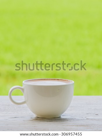 Fresh coffee in a mug shot placed on the left vertical white wooden floor. Behind the green fields blur and poor light.