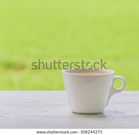 Fresh coffee in a mug shot vertically placed right on the white wood. Behind the green fields blur and poor light.