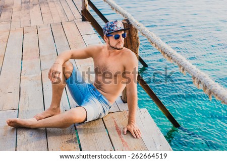 The guy in the denim shorts, sunglasses and a cap sitting on the pier against the sea and watching the sunset