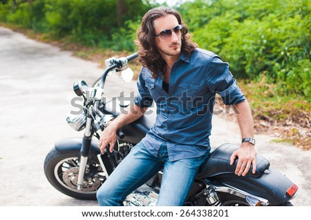 Long-haired brunette guy in sunglasses jeans and a denim shirt posing on a black custom motorcycle