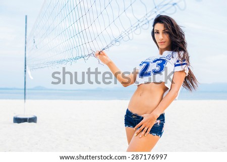 sports sexy brunette in denim shorts and a short t-shirt posing on the volleyball court with net and ball