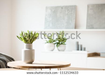 Flores frescas em vaso branco colocado em uma pequena mesa no interior da sala luminosa com pinturas, vasos de plantas e velas nas prateleiras em fundo desfocado