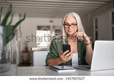 Photo of Happy senior woman using mobile phone while working at home with laptop. Smiling cool old woman wearing eyeglasses messaging with smartphone. Beautiful stylish elderly lady browsing site on cellphone.