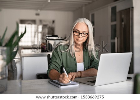 Photo of Senior stylish woman taking notes in notebook while using laptop at home. Old freelancer writing details on book while working on laptop in living room. Focused cool lady writing notary in notepad.